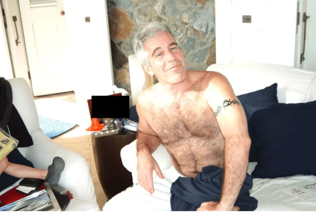 A shirtless Jeffrey Epstein sits on a bed. A young girl is off to his right. (Photo: DOJ)